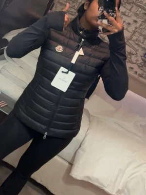 Svart dunväst från Moncler - Snygg svart dunväst från Moncler med hög krage och dragkedja framtill. Västen har det klassiska Moncler-logotypen på bröstet och quiltad design som ger en clean och modern look. Perfekt att slänga över en hoodie eller tröja för extra värme. OBS ÄKTA!😇
