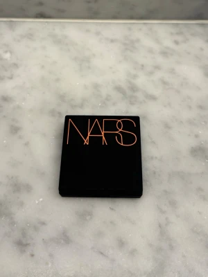 NARS bronzer i mini size - Snygg bronzer från NARS i en stilren svart kompakt med spegel. Pudret har en varm, gyllenbrun ton med lätt skimmer som ger huden en solkysst look. Obs, produkten är i mini size så den är perfekt om du önskar testa den här bronzern eller för att ha i väskan. Helt oanvänd 🩷