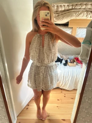 Beige kort klänning med leopardmönster - Säljer en luftig och somrig kort klänning i beige med diskret leopardmönster. Klänningen har halterneck, är ärmlös och har en lös passform med resår i midjan. Perfekt för varma dagar och ger en avslappnad men trendig look. Passar mig som brukar bära S