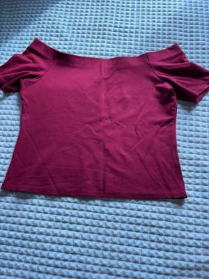Vinröd offshoulder topp - Säljer en vinröd offshoulder topp i mjukt stretchigt material. Toppen har korta ärmar och en figurnära passform som framhäver axlarna. Perfekt att styla med jeans eller kjol för en trendig look. Den är i storlek L men ser även snygg ut på S/M. 