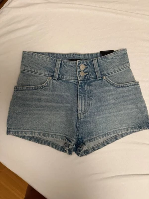 Högmidjade blå jeansshorts XS - Säljer ett par högmidjade blå jeansshorts i klassisk denim med tre knappar framtill och fickor både fram och bak. Shortsen har en snygg, tight passform och är perfekta för varma dagar. Stilsäkra och enkla att matcha med olika toppar.