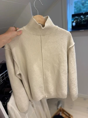 Beige stickad polotröja - Säljer en beige stickad polotröja med hög krage och avslappnad passform. Tröjan har lång ärm och en snygg söm framtill som ger en modern touch. Perfekt att styla med jeans eller kjol för en enkel men trendig look.