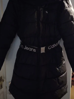 Svart lång dunjacka Calvin Klein - Snygg svart lång dunjacka från Calvin Klein med bälte i midjan där det står 'Calvin Jeans'. Jackan har quiltad design, dragkedja framtill och hög krage. Perfekt för kalla vinterdagar och ger en stilren look.