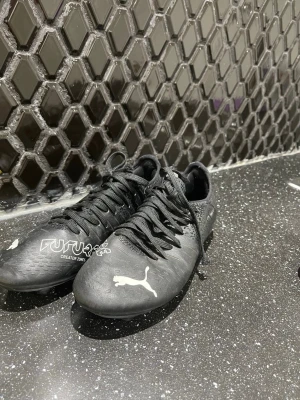 Svarta Puma Future fotbollsskor - Säljer ett par svarta Puma Future fotbollsskor med vita detaljer och logga. Skorna har snörning och låg profil, tillverkade i syntetmaterial för bra grepp och kontroll på planen. Snygg och modern design som passar perfekt för match och träning.
