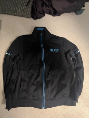 Svart jacka från BOSS med blå detaljer - Snygg svart jacka från BOSS med blå dragkedja och blå logga på bröstet. Jackan har hög krage och ribbade muddar nedtill. Materialet ser ut att vara mjukt och bekvämt, perfekt för lager-på-lager under kyliga dagar.