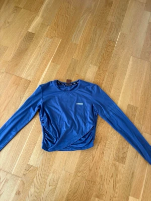 Blå långärmad topp från STRONGER - Snygg blå långärmad topp från STRONGER med rund halsringning och diskret logga på bröstet. Toppen har ett coolt rutmönster i mesh på ena ärmen och är tillverkad i ett stretchigt syntetmaterial. Perfekt för dig som gillar sportig stil. Den är omlott i slutet på tröjan 