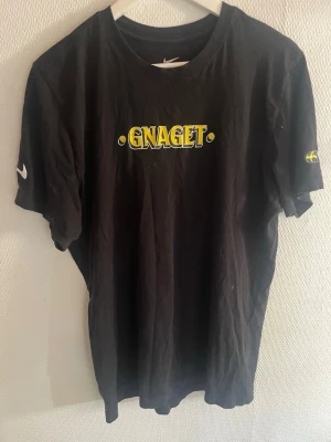 Svart GNAGET t-shirt från Nike - Svart t-shirt från Nike med GNAGET-tryck i gult och vitt på bröstet. På ena ärmen finns en svart och gul flagga och på den andra en Nike logga. Klassisk passform och rund halsringning. Säljer för att den inte kommit till särskilt mycket användning för mig skriv om du har frågor eller undrar något om plagget. Priset går att diskuteras! Nypris 399