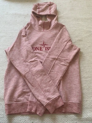 Rosa stone island hoodie med tryck och huva - Säljer en rosa stone island hoodie med tryck framtill och en stor huva. Tröjan har långa ärmar, ribbade muddar och är gjord i ett mjukt sweatshirtmaterial. Perfekt för dig som gillar en avslappnad och snygg stil.