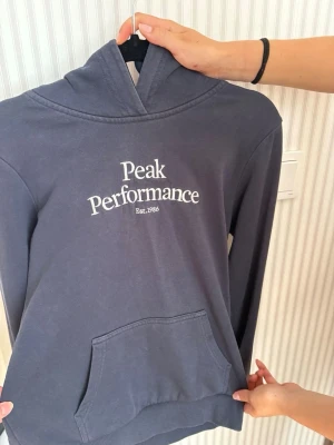 Mörkblå hoodie Peak Performance - Snygg mörkblå hoodie från Peak Performance med vit logga och text på bröstet. Klassisk känguruficka framtill och huva. Perfekt för chill dagar eller när du vill ha en clean och sportig look.