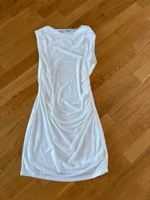 Vit klänning med scrunch - Snygg vit klänning med scrunch på ena sidan och draperade detaljer längs sidorna. Klänningen är ärmlös och har en mjuk, följsam passform som ger en stilren och modern look.
