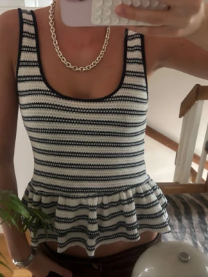 Randig peplum linnetopp - Supersöt randig linnetopp med peplumvolang nertill. Toppen är ärmlös och har breda axelband samt rundad urringning. Materialet är stickat i vitt och mörkblått för en fräsch look. Perfekt att styla med jeans eller kjol.