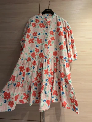 Blommig klänning - Klänning med färgglatt blommönster i rött, blått, rosa och grönt på vit botten. Klänningen har 6 knappar framtill och resår vid ärmslut. Oanvänd men tyvärr bortklippta etiketter.