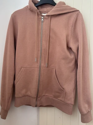 Rosa zip hoodie med huva - Säljer en mjuk rosa hoodie med dragkedja framtill och stor ficka på magen. Tröjan har huva med dragsko och ribbade muddar vid ärmar och nederkant. Perfekt för en avslappnad och chill stil.