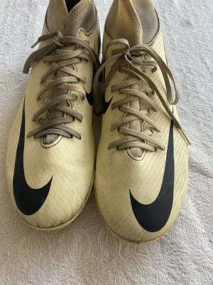 Beigea Nike fotbollsskor med dobbar - Säljer ett par beigea Nike fotbollsskor med svarta detaljer och stora Nike-loggor på ovansidan. Skorna har snörning och slitstarka dobbar för bra grepp på planen. Tillverkade i syntetmaterial som ger en lätt känsla och bra bollkontroll.