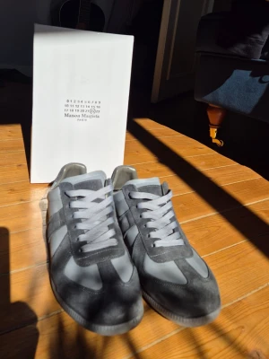 Maison Margiela grå sneakers - Maison Margiela sneakers i grått med vita detaljer och snörning. Skorna har en klassisk låg siluett och är tillverkade i en mix av mocka och läder. Snygg retrostil som passar till många olika outfits.