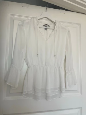 Vit blus med volangärm från H&M - Säljer en superfin vit blus från H&M med v-ringning och tunna knytband. Blusen har långa volangärmar och smock i midjan för en snygg siluett. Tillverkad i ett lätt, luftigt tyg som ger en romantisk känsla. Säljer då den tyvärr aldrig kommit till användning.