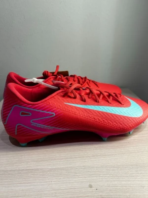 Nike Mercurial röda fotbollsskor - Snygga röda Nike Mercurial fotbollsskor med turkosa detaljer och logga. Skorna har låg profil, snörning och dobbar för bra grepp på planen. Tillverkade i syntetmaterial för lätt vikt och bra bollkontroll. Perfekta för dig som vill sticka ut på fotbollsplanen.