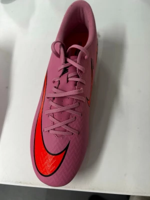 Rosa Nike fotbollsskor med dobbar - Säljer ett par snygga rosa Nike fotbollsskor med röda och svarta detaljer. Skorna har klassisk snörning, låg profil och dobbar för bra grepp på planen. Materialet är syntetiskt och designen är sportig med stora Nike-loggor på sidorna.