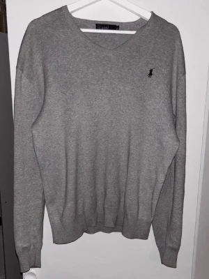 Grå V-ringad tröja från Polo Ralph Lauren - Klassisk grå V-ringad tröja från Polo Ralph Lauren med diskret broderad logga på bröstet. Tröjan har långa ärmar och ribbade muddar vid ärmslut och nederkant. Perfekt att bära över en skjorta eller t-shirt.