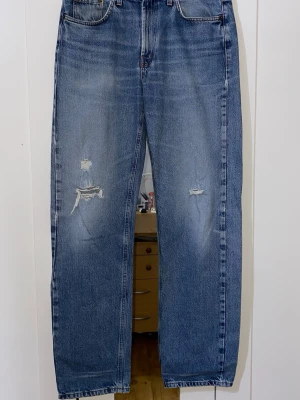 Blå raka jeans med slitningar - Säljer ett par blå raka jeans med slitningar på båda benen och klassisk femficksdesign. Jeansen har hög midja och är tillverkade i denim med en snygg tvättad look. Perfekta för en avslappnad och trendig stil.