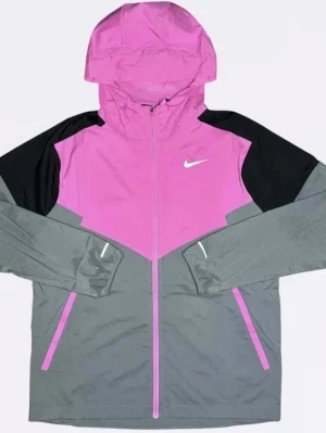 Nike vindjacka rosa och grå - Snygg vindjacka från Nike i rosa, grått och svart. Jackan har huva, hel dragkedja framtill och två fickor med dragkedja. Ikonisk Nike-logga på bröstet. Perfekt för blåsiga dagar och sportiga outfits.