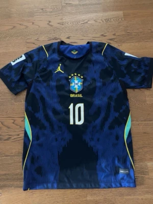 Blå Brasilien matchtröja nummer 10 - Snygg blå matchtröja från Brasilien med nummer 10 på bröstet och CBF-logga. Tröjan har korta ärmar, gula detaljer och ett sportigt mönster i blått och svart. Perfekt för fotbollsträning eller att bära till vardags. Med neymar tryck på ryggen