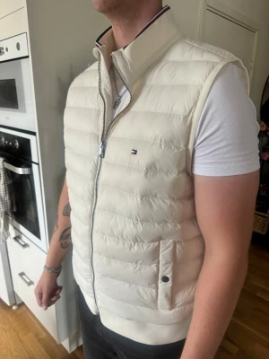 Vit dunväst från Tommy Hilfiger - Snygg vit dunväst från Tommy Hilfiger (ÄKTA) med diskret logga på bröstet. Västen har ståkrage, dragkedja framtill och två fickor med knappstängning. Perfekt att slänga över en hoodie eller t-shirt för en clean och stilren look. Endast använd 2 gånger!!🤍