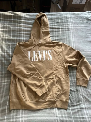Beige hoodie från Levi's med tryck - Säljer en beige hoodie från Levi's i storlek L. Hoodien har stor vit Levi's-logga på bröstet, känguruficka och ribbade muddar. Tillverkad i mjukt bomullsmaterial och har en relaxed fit. Perfekt för en chill och avslappnad stil.