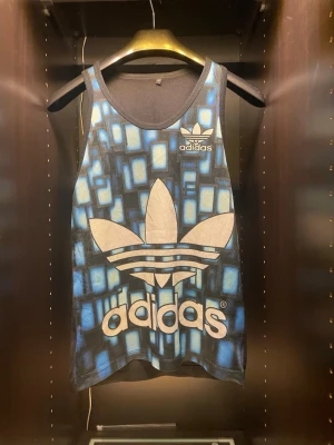 Blåmönstrad linnetopp från Adidas - Snygg linnetopp från Adidas med stort vitt logotryck framtill och blått geometriskt mönster. Modellen är ärmlös och har rund halsringning. Perfekt för dig som vill sticka ut med sportig stil.