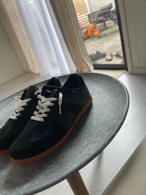Svarta sneakers med vit sula - Snygga svarta sneakers med vita skosnören och brun gummisula. Skorna har en klassisk låg profil och är tillverkade i en mix av mocka och textil. Perfekta för dig som gillar stilrena och tidlösa sneakers.