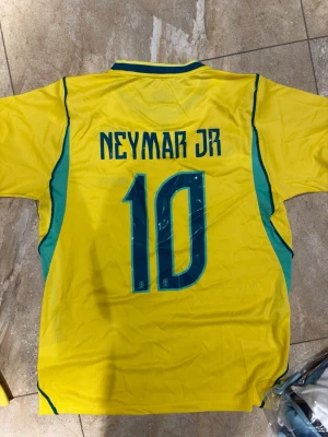 Brasilien Neymar JR matchtröja gul - Säljer en gul Brasilien fotbollströja från Nike med Neymar JR och nummer 10 på ryggen. Tröjan har gröna detaljer, korta ärmar och CBF-emblemet på bröstet. Perfekt för dig som älskar fotboll och vill stötta Brasilien på match!