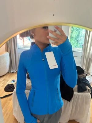 Blå ziptröja från Lululemon - Snygg blå ziptröja från Lululemon med figurnära passform och hög krage. Tröjan har tumhål vid ärmsluten, två dragkedjeförsedda fickor och är tillverkad i ett stretchigt funktionsmaterial. Perfekt för träning eller som lager på lager.