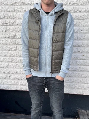 Olivgrön väst - Snygg olivgrön dunväst med dragkedja framtill. Västen har en klassisk, rak passform och är perfekt att slänga över en hoodie för en avslappnad look. Tillverkad i ett glansigt material som ger en modern känsla.
