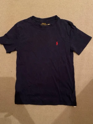 Marinblå t-shirt från Polo Ralph Lauren - Klassisk marinblå t-shirt från Polo Ralph Lauren med röd broderad logga på bröstet. T-shirten har rund halsringning och korta ärmar. Tillverkad i mjuk bomull för en skön känsla och enkel stil. Den har inga hål och är i väldigt fint skick. Tveka inte att höra av dig.                 Mvh Sam