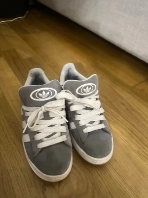 Gråa Adidas sneakers med vita detaljer - Snygga gråa Adidas sneakers med vita ränder och breda vita skosnören. Skorna har en låg profil och är tillverkade i mockaliknande material. Adidas-loggan syns tydligt på plösen. Perfekta för en avslappnad streetwear-look. De har ett litet hål på sidan som man kan se på sista bilden 