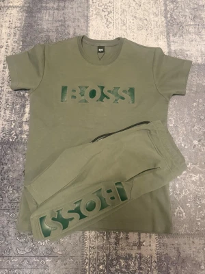 Grönt loungewear-set från BOSS - Snyggt loungewear-set i olivgrönt från BOSS med matchande t-shirt och byxor. Båda plaggen har ett stort BOSS-tryck i grönt framtill. T-shirten har rund hals och byxorna har resår i midjan samt muddar vid bensluten. Perfekt för chill dagar hemma.