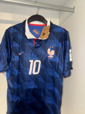 Frankrike fotbollströja Mbappé #10 - Snygg blå fotbollströja från Nike med Frankrikes landslagsmärke och nummer 10, Mbappé på ryggen. Tröjan har korta ärmar, vit krage med röd detalj och ett mönster i olika blå nyanser. 