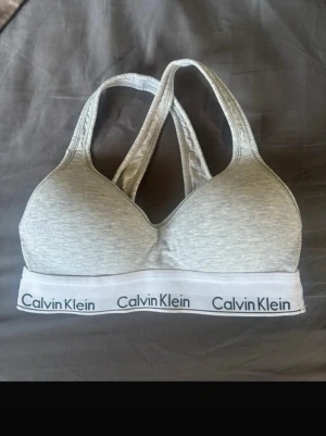 Calvin Klein bh  - Sportig grå bh från Calvin Klein med vadderade kupor och bred resår med logga runt om. Bh:n har brottarrygg och är tillverkad i mjukt stretchigt material för extra komfort. Perfekt för en avslappnad och stilren look.