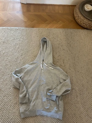 Ljusgrå zip hoodie med huva - En enkel och stilren ljusgrå hoodie med dragkedja framtill och justerbar huva med vita snören. Hoodien har långa ärmar, ribbade muddar och två fickor framtill. Perfekt för en avslappnad look.