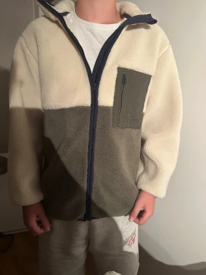 Teddy jacka med dragkedja - Mysig fleecejacka i beige och olivgrönt med hel dragkedja framtill och hög krage. Jackan har en kontrasterande bröstficka med dragkedja och mörkblå detaljer längs dragkedjan. Perfekt för kyliga dagar och enkel att matcha med olika outfits.