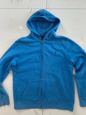 Blå hoodie från Polo Ralph Lauren - Snygg blå hoodie från Polo Ralph Lauren med dragkedja och luva. Hoodien köptes i storlek Xl på barnavdelningen men var till får 18/20 år skulle säga att den även passar vanlig storlek S och M