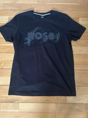 Svart BOSS t-shirt med tryck - Snygg svart t-shirt från BOSS med ett stort, diskret tryck på bröstet. Klassisk rund halsringning och korta ärmar. Tillverkad i mjuk bomull som känns skön mot huden. Perfekt för en clean och stilren look.