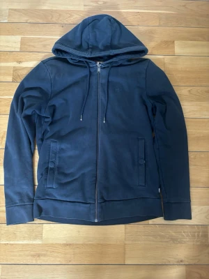Mörkblå zip hoodie med huva från Boss - Säljer en mörkblå hoodie med dragkedja framtill och två fickor med knappstängning. Tröjan har justerbar huva med snören och ribbade muddar vid ärmslut och nederkant. Perfekt för en avslappnad och stilren look.