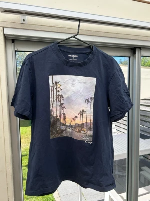 Mörkblå t-shirt med palmtryck - Snygg mörkblå t-shirt med ett stort tryck av palmer och solnedgång på framsidan. T-shirten har rund hals och korta ärmar. Perfekt för dig som gillar avslappnad stil och coola motiv.