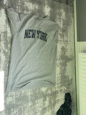 Grå t-shirt med New York-tryck - Säljer en grå t-shirt med svart text 'NEW YORK' på bröstet. Klassisk rund hals och korta ärmar. Perfekt för dig som gillar enkel streetstyle och vill ha en basic tee med coolt tryck.