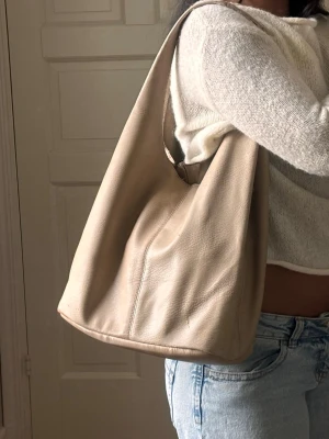 Beige axelväska i mjukt skinn - Stilren beige axelväska i mjukt skinn med minimalistisk design och rymlig insida. Väskan har en enkel, slouchy form och bred axelrem som gör den bekväm att bära. Perfekt för dig som gillar en clean och avslappnad look.