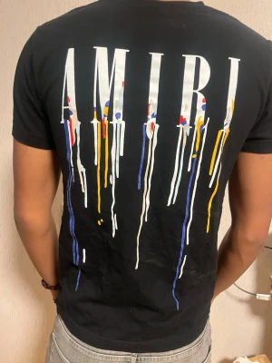 Svart AMIRI t-shirt med färgstänk - Snygg svart t-shirt från AMIRI med stort vitt logotryck på ryggen och färgstänk i blått, gult, rött och vitt som rinner ner från bokstäverna. T-shirten har rund hals och korta ärmar. Perfekt för dig som gillar streetwear och unika detaljer.