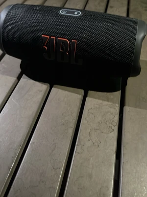 JBL charge 5 svart trådlös högtalare - Säljer en svart trådlös högtalare från JBL med robust cylinderformad design. Högtalaren har ett slitstarkt tygmaterial och tydlig JBL-logga i orange. Utrustad med USB-C laddningsport och gummidetaljer för extra grepp. OBS ingår dock inte i orginal förpackning 