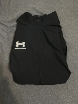 Svart träningsjacka Under Armour - Säljer en svart träningsjacka från Under Armour med dragkedja framtill och logga i vitt på bröstet. Jackan har hög krage och är tillverkad i ett lätt, stretchigt material som passar perfekt till träning och sportaktiviteter.