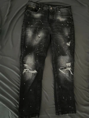 Svarta distressed jeans Supply & Demand - Säljer ett par svarta jeans Har änvämt dem en gång ungefär eller två från Supply & Demand med slitningar och färgstänk för en edgy look. Jeansen har raka ben, klassisk femficksmodell och coola detaljer som trasiga knän och blekta partier. Perfekta för dig som gillar streetstyle.